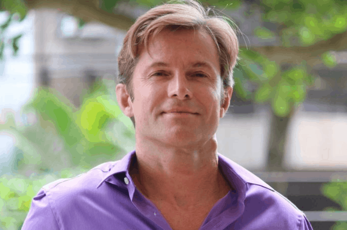 Claudio Heinrich hoje, vestindo um quimono branco de Jiu-jitsu com a faixa preta amarrada na cintura, sorrindo de forma tranquila enquanto segura o filho Karl no colo.