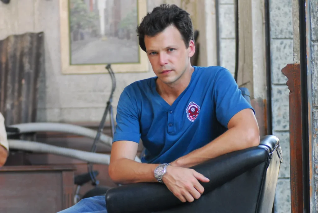 Um retrato de Guilherme Berenguer hoje em um set de filmagem em Los Angeles. Ele est&aacute; usando um bon&eacute; escuro, fones de ouvido profissionais no pesco&ccedil;o e observa atentamente um monitor de v&iacute;deo enquanto segura uma prancheta.