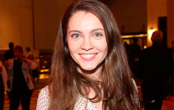 m retrato contempor&acirc;neo de Cec&iacute;lia Dassi em seu consult&oacute;rio, sentada em uma poltrona de tecido cinza com uma estante de livros de psicologia ao fundo.