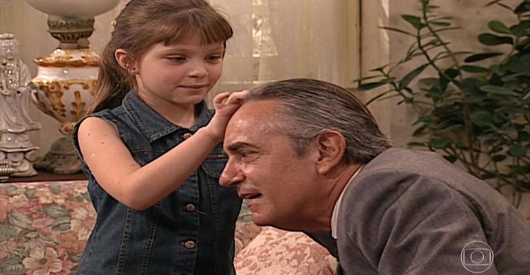 m retrato contempor&acirc;neo de Cec&iacute;lia Dassi em seu consult&oacute;rio, sentada em uma poltrona de tecido cinza com uma estante de livros de psicologia ao fundo.