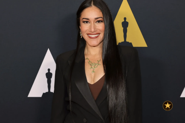 Plano médio de Q'orianka Kilcher em uma coletiva de imprensa com expressão séria e olhar direto para as lentes. Uma luz fria recorta o rosto da atriz